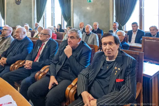 Grupa mężczyzn siedzących w sali, w formalnych strojach, podczas spotkania lub wydarzenia.