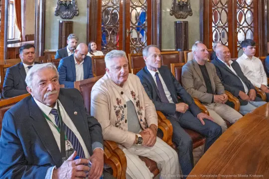Grupa osób siedzących w sali, w formalnych strojach, podczas spotkania.