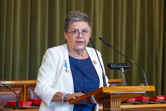 Kobieta w okularach, przemawiająca z mównicy, ubrana w białą marynarkę i granatową bluzkę.