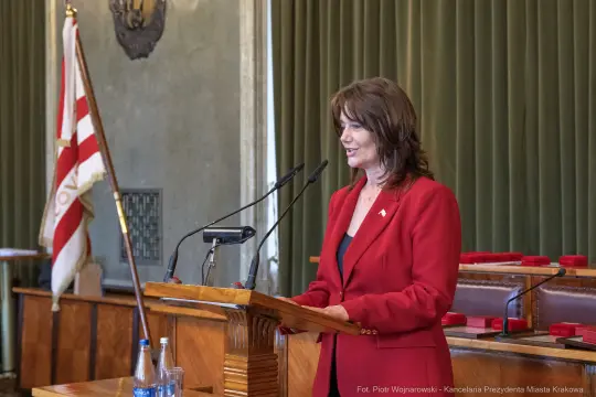 Kobieta w czerwonym żakiecie przemawia przy mównicy w sali obrad.
