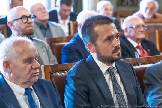 Mężczyźni w garniturach siedzący w sali obrad, skupieni na dyskusji.
