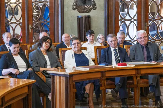 Grupa osób siedzących w sali, w tle dekoracyjne okna i stoły.