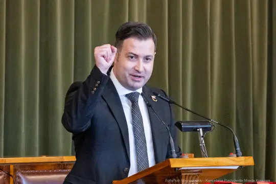 Mężczyzna w garniturze podnosi rękę w geście przemówienia.