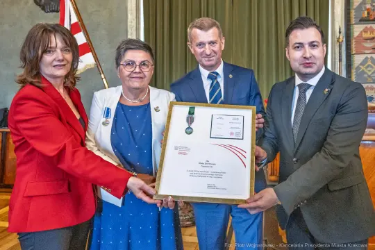 Wręczenie medalu i dyplomu w uroczystej atmosferze, cztery osoby trzymają ramkę z wyróżnieniem.