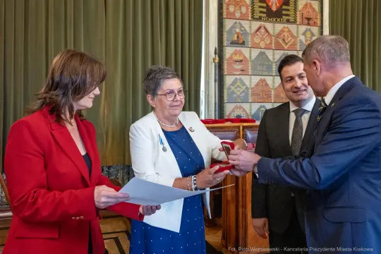 Wręczenie odznaczenia w uroczystej atmosferze, cztery osoby w formalnych strojach.