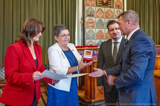 Wręczenie nagrody w sali obrad, cztery osoby w formalnych strojach, uśmiechy i gratulacje.