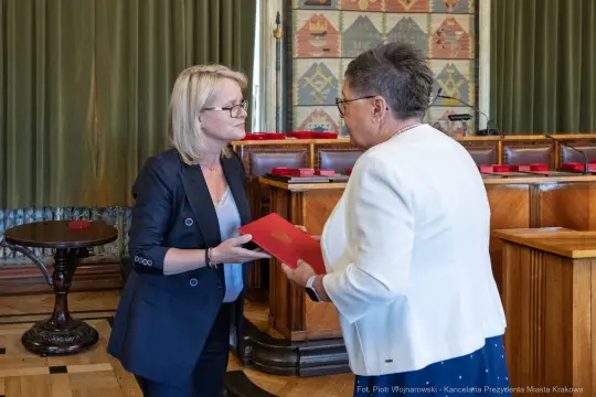 Wręczenie dokumentów w sali obrad, dwie kobiety w formalnych strojach.