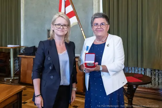 Dwie kobiety w eleganckich strojach, jedna z medalem w dłoni, w sali z dekoracjami.