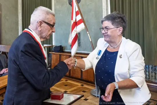 Wręczenie odznaczenia przez kobietę mężczyźnie w formalnym otoczeniu.