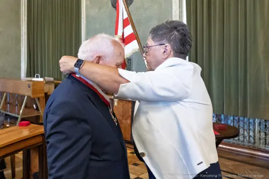 Wręczenie odznaczenia przez kobietę mężczyźnie w sali z flagą.