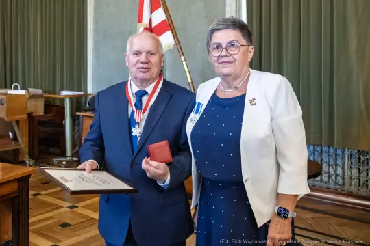 Mężczyzna z odznaczeniem i kobietą w eleganckim stroju, trzymający dyplom w sali.