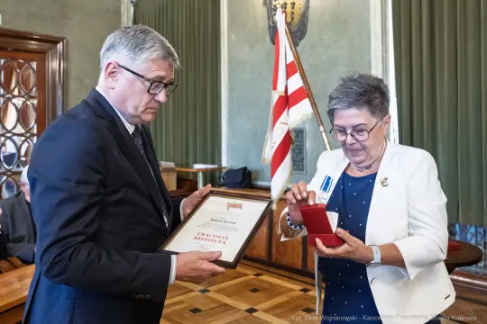 Wręczenie odznaczenia przez kobietę mężczyźnie w eleganckim otoczeniu.