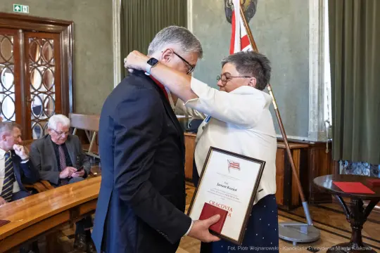 Wręczenie nagrody przez kobietę mężczyźnie w sali z flagą i dokumentami.