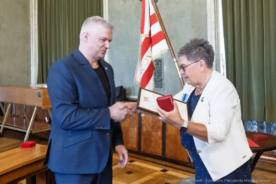 Wręczenie odznaczenia w sali obrad, mężczyzna i kobieta w formalnych strojach.
