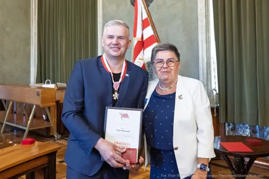 Mężczyzna z medalem i dyplomem stoi obok kobiety w okularach, oboje uśmiechnięci.