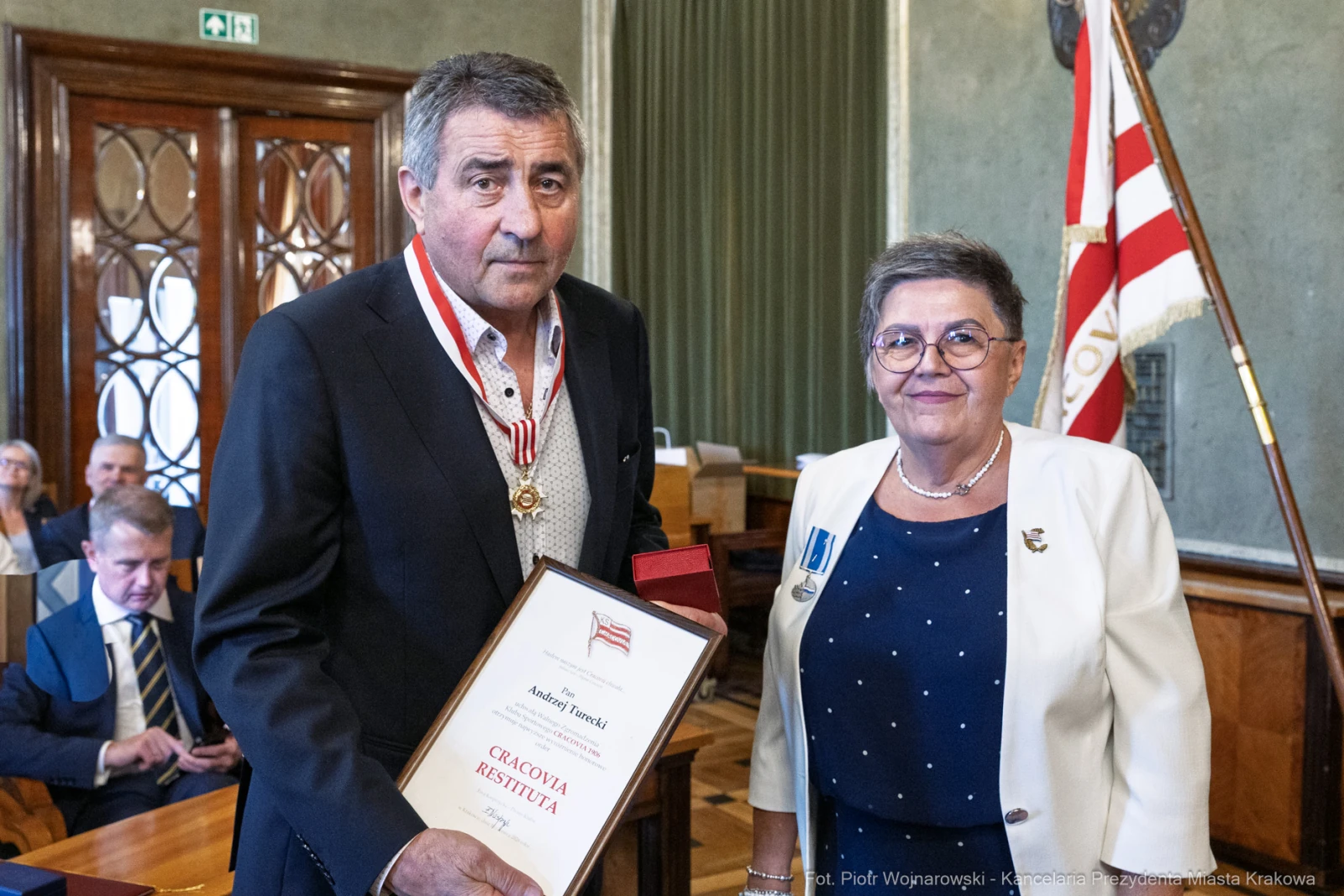 Mężczyzna z medalem i dyplomem, obok kobieta w białej marynarce, w tle sala z flagą.  Autor: Piotr Wojnarowski