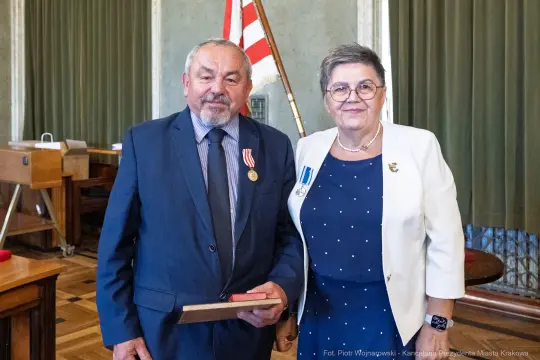 Mężczyzna z medalem i kobieta z odznaczeniem, oboje w formalnych strojach, w sali z flagą.