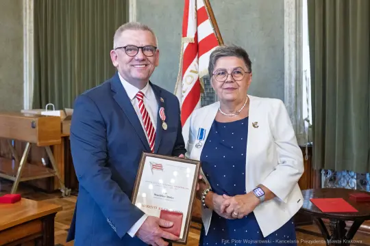 Mężczyzna i kobieta wręczają sobie dyplom w sali z flagą.
