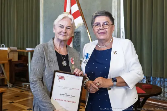Dwie kobiety trzymające dyplom z wyróżnieniem, jedna z odznaczeniem na piersi.