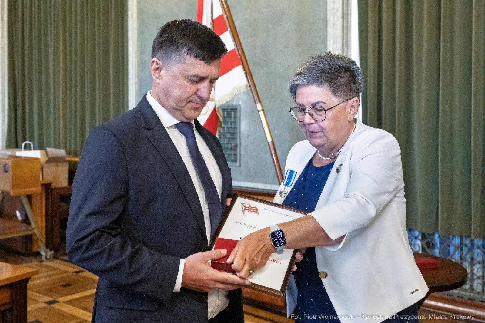 Wręczenie odznaczenia przez kobietę mężczyźnie w sali z flagą.  Autor: Piotr Wojnarowski