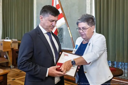 Wręczenie odznaczenia przez kobietę mężczyźnie w sali z flagą.