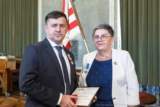 Mężczyzna i kobieta trzymają dyplom w sali z flagami.
