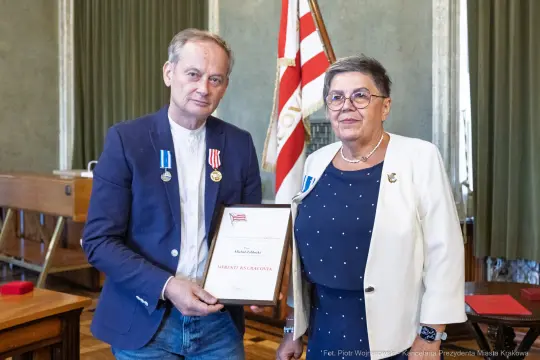 Mężczyzna i kobieta z medalami, wręczający sobie dyplom w sali z flagą.