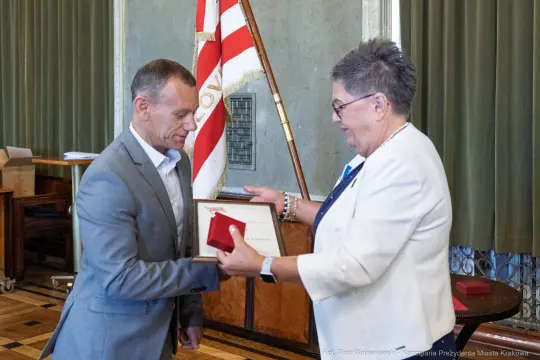 Wręczenie nagrody w sali z flagą i dyplomem.
