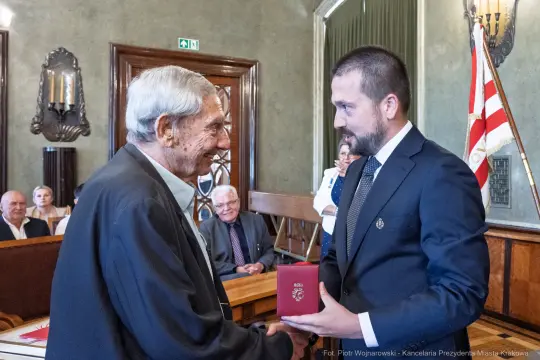 Wręczenie odznaczenia przez mężczyznę w garniturze starszemu mężczyźnie w sali obrad.