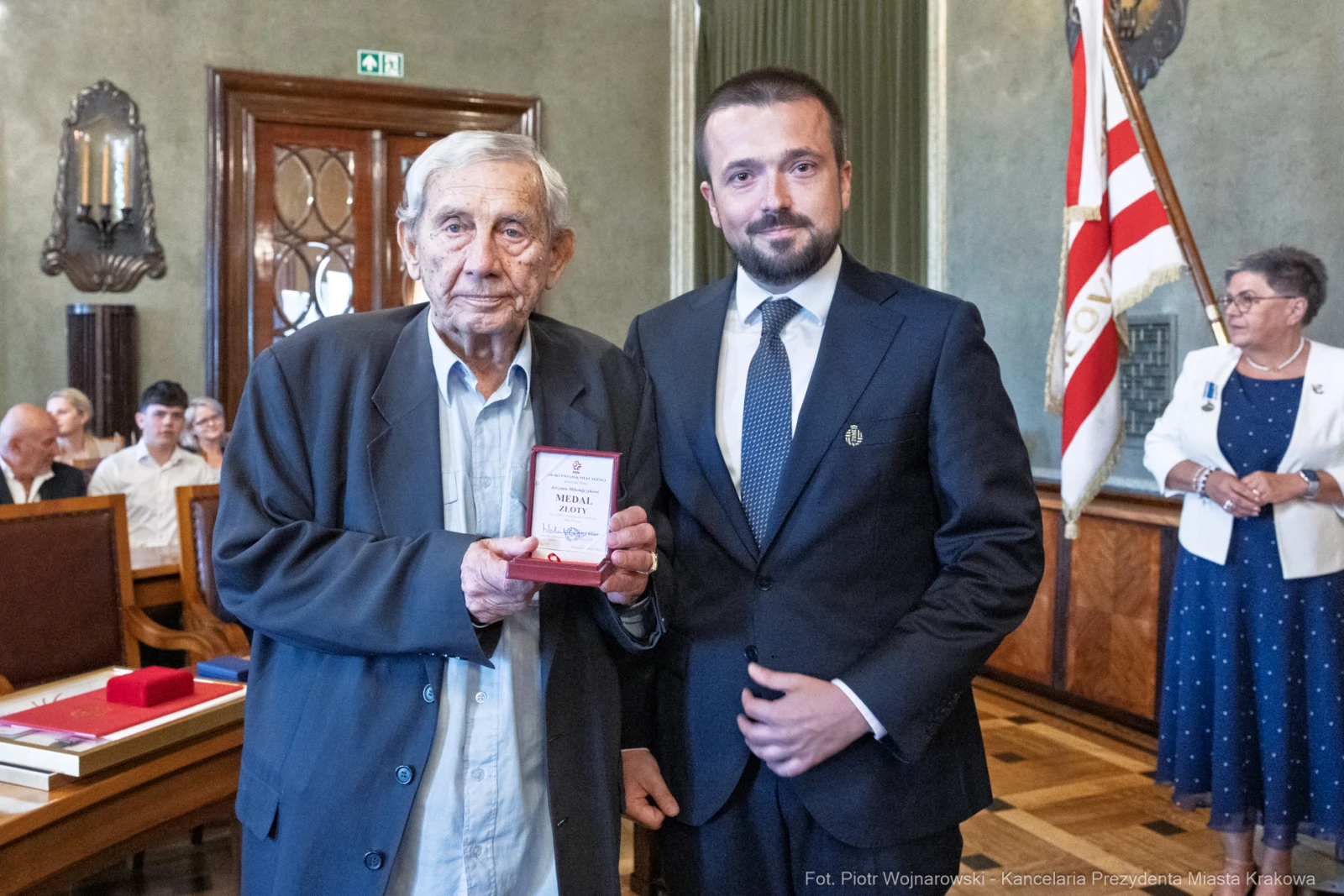 Mężczyzna wręcza medal starszemu mężczyźnie w eleganckim wnętrzu z gośćmi w tle.  Autor: Piotr Wojnarowski