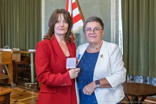 Wręczenie odznaczenia przez jedną kobietę drugiej w eleganckim otoczeniu.