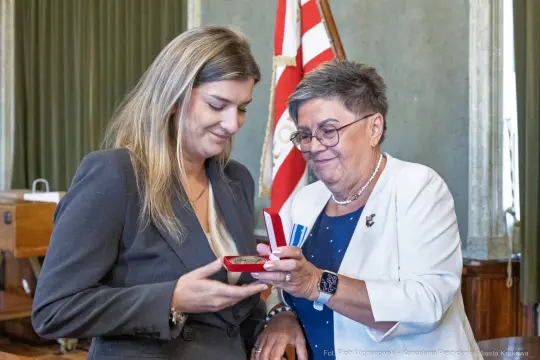 Wręczenie medalu przez kobietę w okularach drugiej kobiecie w garniturze.