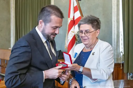 Wręczenie medalu przez kobietę w okularach mężczyźnie w garniturze w sali z flagami.
