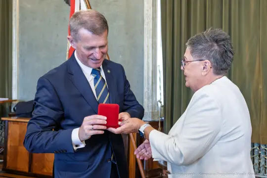 Wręczenie odznaczenia przez mężczyznę kobiecie w oficjalnym otoczeniu.