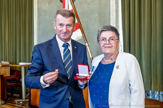Wręczenie odznaczenia przez mężczyznę i kobietę w oficjalnych strojach.