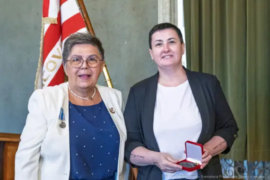 Dwie kobiety z medalem w eleganckich strojach, jedna z nich uśmiechnięta.