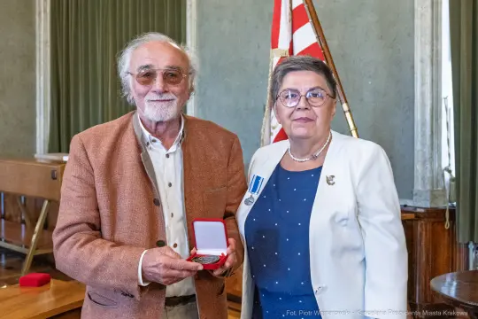 Mężczyzna w okularach wręcza medal kobiecie w garniturze, oboje uśmiechnięci.