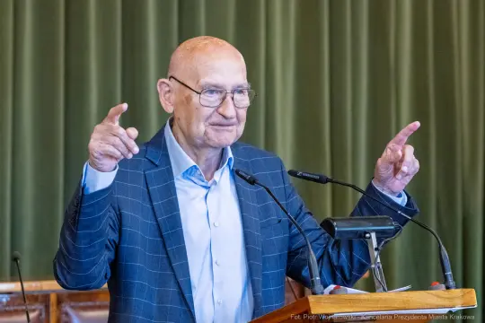 Mężczyzna w okularach wygłasza przemówienie, gestykulując rękami.