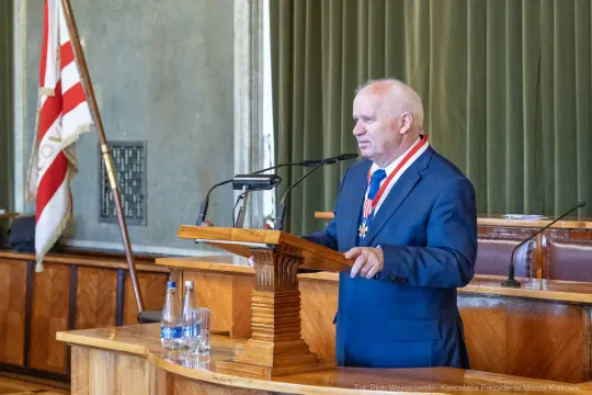 Mężczyzna w garniturze z medalem, przemawiający z mównicy w sali obrad.