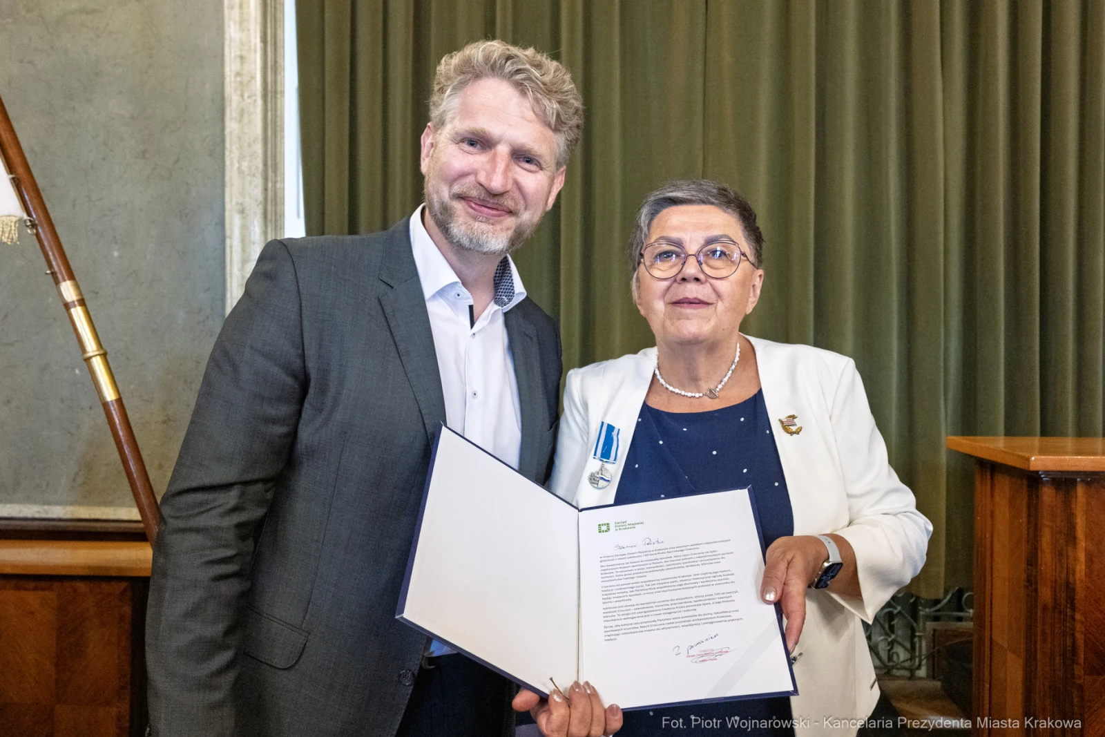 Mężczyzna i kobieta trzymają dokument, oboje uśmiechnięci, w tle zasłony i drewniana ściana.  Autor: Piotr Wojnarowski