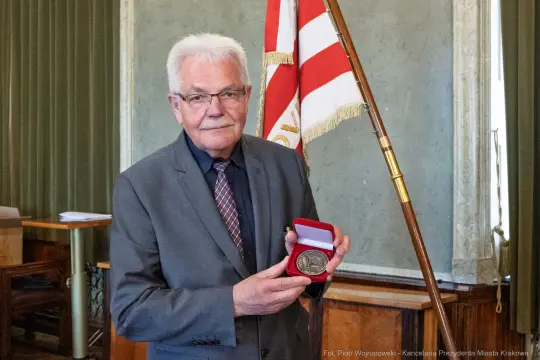 Mężczyzna w okularach trzyma medal w czerwonym etui, stoi obok flagi.