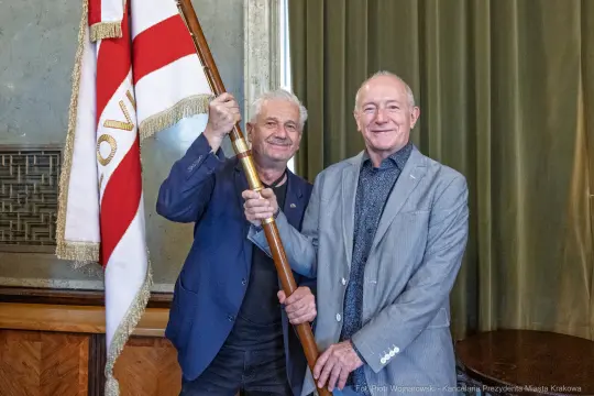 Dwaj mężczyźni trzymają sztandar w sali z zielonymi zasłonami.