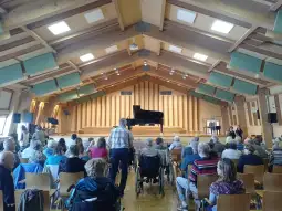 Sala koncertowa z fortepianem, publiczność siedząca na krzesłach, atmosfera oczekiwania.