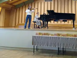Młoda skrzypaczka gra na scenie, obok niej siedzi pianistka. Na stole leżą słodycze.