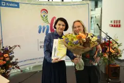 Dwie kobiety trzymają bukiet kwiatów i książkę, w tle dekoracje z logo Krakowa.