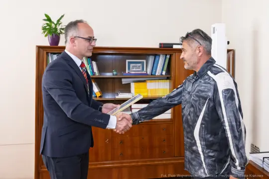 Mężczyźni w garniturze i skórzanej kurtce podają sobie ręce, wręczając dokument.