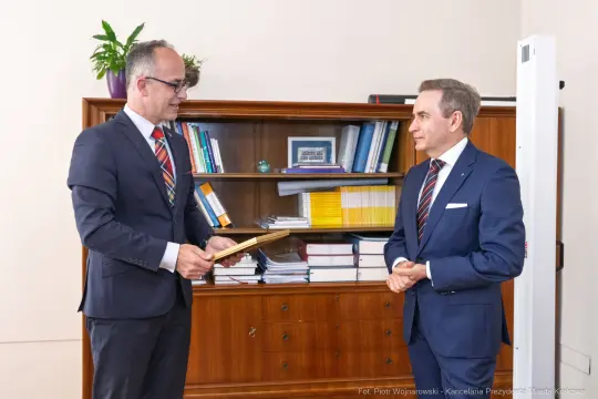 Dwaj mężczyźni w garniturach, jeden wręcza dokument drugiemu w biurowym otoczeniu.