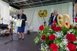 Kobieta przemawia z mikrofonem, obok niej stoi mężczyzna, w tle dekoracje z cyfrą 60 i bukiet róż.