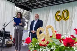 Kobieta przemawia na uroczystości z okazji 60-lecia, obok mężczyzna, w tle złote balony z cyfrą 60.