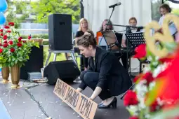 Kobieta przygotowuje banner na scenie, w tle muzycy i dekoracje kwiatowe.